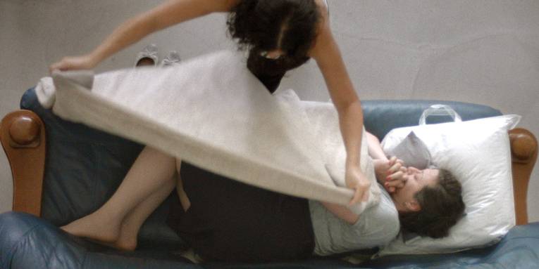 Eine Frau liegt schlafend auf einem Sofa - eine gefüllte Plastiktüte hat sie als Kopfkissen. Eine weitere Frau deckt sie zu.